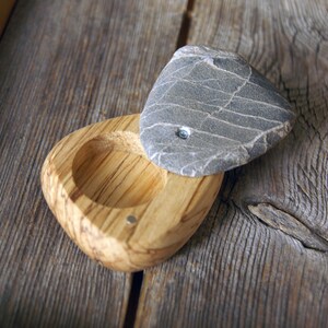 May include: A handcrafted wooden bowl with a gray stone lid. The bowl is made of light-colored wood with a natural grain pattern. The stone lid is gray with white veining. The bowl and lid are on a weathered wooden surface.