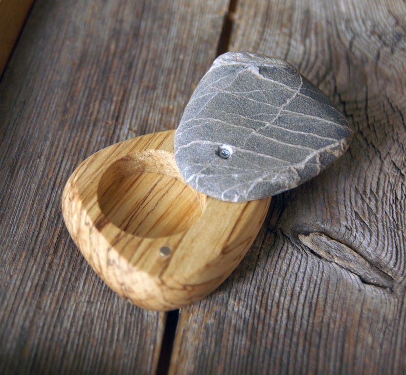 May include: A handcrafted wooden bowl with a gray stone lid. The bowl is made of light-colored wood with a natural grain pattern. The stone lid is gray with white veining. The bowl and lid are on a weathered wooden surface.