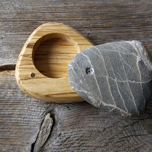 May include: A handmade wooden box with a natural stone lid. The box is made of light-colored wood with a triangular shape and a circular indentation. The lid is a gray stone with a unique pattern, attached with a screw.