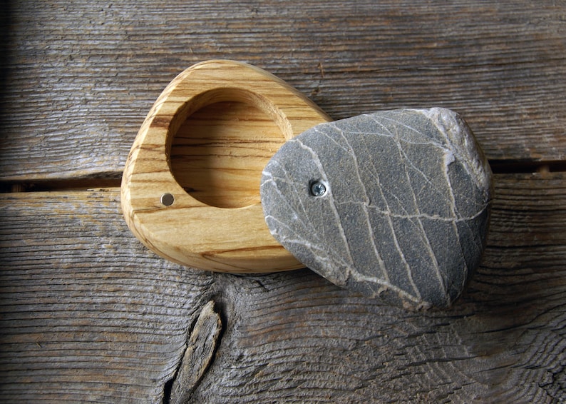 May include: A handmade wooden box with a natural stone lid. The box is made of light-colored wood with a triangular shape and a circular indentation. The lid is a gray stone with a unique pattern, attached with a screw.