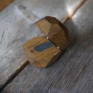 May include: A wooden ring box with a hinged lid. The box is made of light brown wood with a dark brown stain. The lid is open and the inside of the box is lined with a dark gray felt.