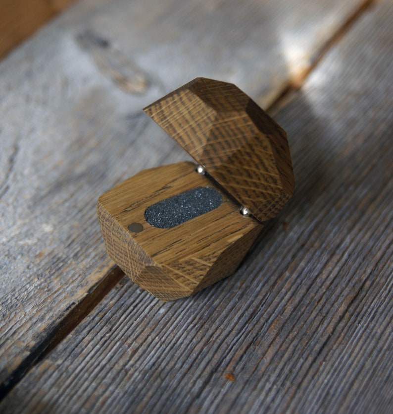 May include: A wooden ring box with a hinged lid. The box is made of light brown wood with a dark brown stain. The lid is open and the inside of the box is lined with a dark gray felt.