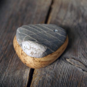 May include: A handcrafted, decorative stone paperweight. The paperweight features a gray stone top with natural veining, set atop a rounded, light-brown wooden base. The design showcases a contrast of textures and colors.