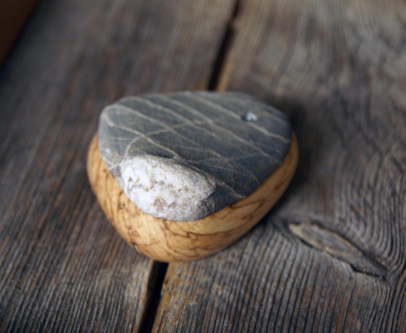 May include: A handcrafted, decorative stone paperweight. The paperweight features a gray stone top with natural veining, set atop a rounded, light-brown wooden base. The design showcases a contrast of textures and colors.