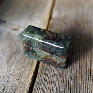 May include: A rectangular lighter case featuring a polished finish with a marbled green resin body embedded with dark brown pine cone fragments. The object has a smooth, glossy surface and a distinct horizontal seam separating the lid from the base.