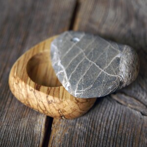 May include: A small, handcrafted wooden box with a natural wood grain finish. The box is oval-shaped with a circular indentation. A gray, heart-shaped stone serves as a lid, adding a unique, natural element to the design.