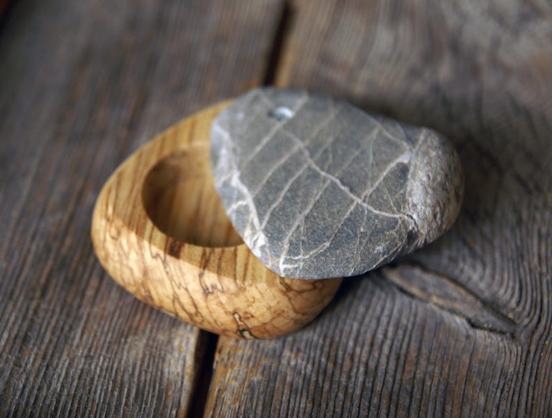 May include: A small, handcrafted wooden box with a natural wood grain finish. The box is oval-shaped with a circular indentation. A gray, heart-shaped stone serves as a lid, adding a unique, natural element to the design.