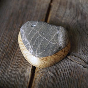 May include: A gray stone with white veining sits atop a light-colored wooden base. The stone has a small hole drilled into it. The wooden base has a curved shape and a natural wood grain pattern. The items are displayed on a weathered wooden surface.