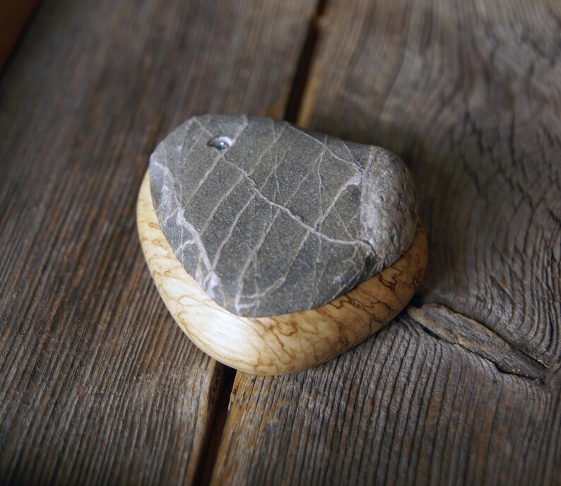May include: A gray stone with white veining sits atop a light-colored wooden base. The stone has a small hole drilled into it. The wooden base has a curved shape and a natural wood grain pattern. The items are displayed on a weathered wooden surface.