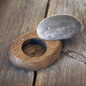 May include: A small, oval-shaped wooden container with a hollow centre and a smooth, grey, rounded stone lid. The lid is attached to the wooden base with a small metal hinge, allowing it to pivot open to reveal the interior compartment.