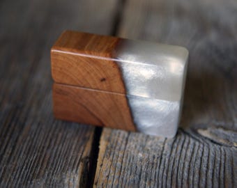 May include: A small, rectangular wooden box with a hinged lid. The box is split diagonally, with one half of the box made of polished, reddish-brown wood and the other half of a shimmering, pearlescent silver material. The box is resting on a weathered wooden surface.