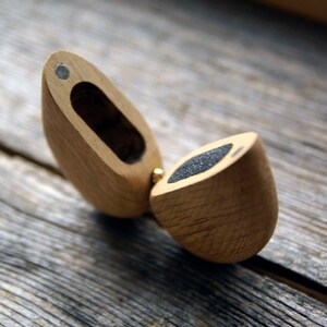 May include: A wooden ring box with a hinged lid. The box is made of light brown wood and has a dark gray felt lining. The box is open and the lid is propped up.
