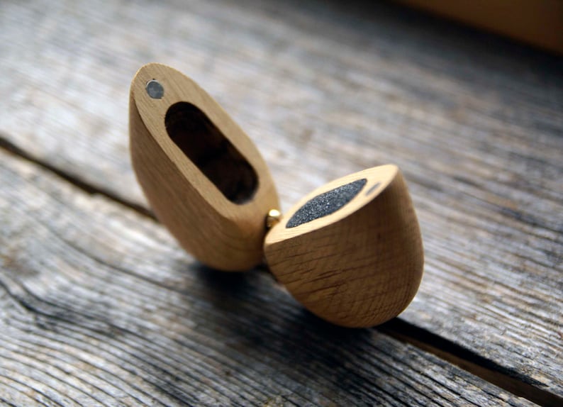 May include: A wooden ring box with a hinged lid. The box is made of light brown wood and has a dark gray felt lining. The box is open and the lid is propped up.