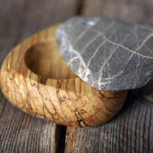 May include: A handcrafted wooden box with a natural stone lid. The box is made of light-colored wood with dark brown grain patterns. The lid is a gray stone with white veining. The box is open, revealing its interior.