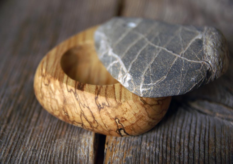 May include: A handcrafted wooden box with a natural stone lid. The box is made of light-colored wood with dark brown grain patterns. The lid is a gray stone with white veining. The box is open, revealing its interior.