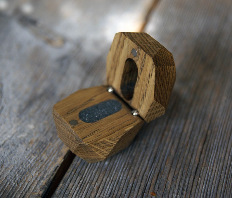 May include: A wooden ring box with a hinged lid, made of light brown oak wood. The box is open, revealing a gray felt lining inside.