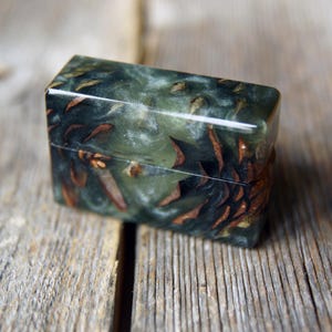 May include: A rectangular lighter case crafted from a combination of dark green resin and natural brown pine cone segments. The surface is smooth and polished, featuring a visible horizontal seam where the lid meets the base of the rectangular object.