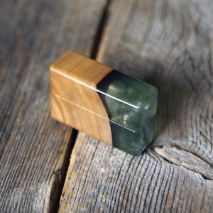 Pode incluir: Um objeto em forma de bloco retangular com uma combinação de madeira castanho-clara de um lado e uma resina verde-azeitona translúcida e semiopaca do outro, unidos por uma costura diagonal que atravessa a superfície da peça retangular.