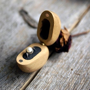 May include: A wooden ring box with a hinged lid, open to reveal a ring with a pearl gemstone. The box is made of light brown wood and has a dark brown velvet interior.
