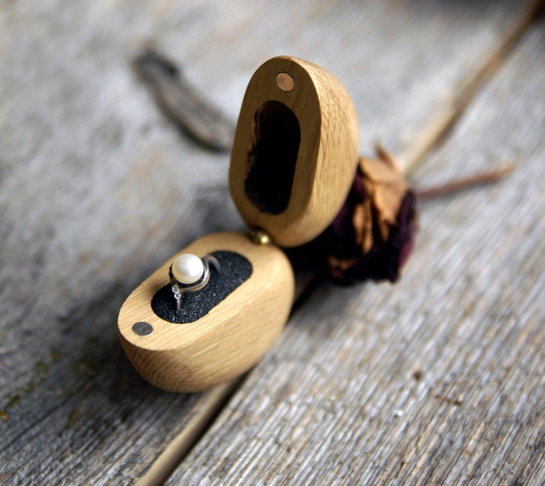 May include: A wooden ring box with a hinged lid, open to reveal a ring with a pearl gemstone. The box is made of light brown wood and has a dark brown velvet interior.