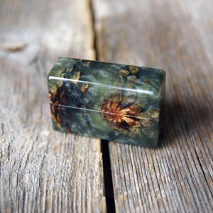 May include: A rectangular box featuring a polished finish with a marbled green and grey resin body. The surface displays embedded natural pine cone segments with visible brown and tan textures, creating a distinct organic pattern across the lid.