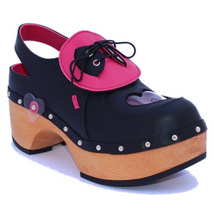 May include: Black and pink platform clog shoe with a heart-shaped cutout and accents. The shoe features a wooden sole, a pink heart-shaped detail, and a black strap. The shoe has a heel height of approximately 4 inches.