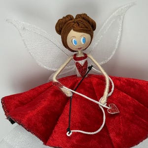 May include: A Valentine's Day fairy doll with a red velvet dress and white tulle underskirt. The doll has brown hair, blue eyes, and white wings. It holds a bow and arrow with a heart-shaped tip.