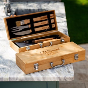 May include: A wooden carrying case with a handle and engraved text "The Breemont" on the top. The case is open to reveal a set of grilling tools, including a spatula, tongs, a fork, and a brush.