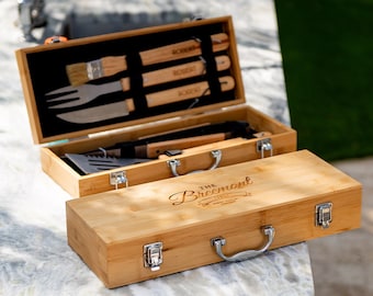 May include: A wooden carrying case with a handle and engraved text "The Breemont" on the top. The case is open to reveal a set of grilling tools, including a spatula, tongs, a fork, and a brush.