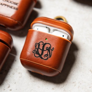 May include: Three brown leather AirPod cases with different engravings. One case says "Choose Happiness", another says "Shiloh", and the third has a monogram.