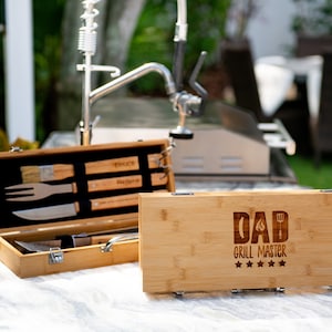 May include: A wooden box with a grill set inside. The box is engraved with the text "DAD Grill Master" and five stars. The box is sitting on a countertop.