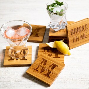 May include: Set of six bamboo coasters with a personalized initial and the name "Marshall" engraved on each. One coaster has the text "Drinks on me" engraved on it. A glass of pink drink with ice cubes and a lemon wedge are on top of the coasters.