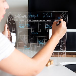 May include: A clear acrylic dry-erase calendar with a black grid and the month of August 2021 written at the top. The calendar is decorated with a leaf design and has handwritten appointments in blue ink.