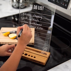 Könnte beinhalten: Ein transparentes Acryl-To-Do-Liste-Brett mit einem Bambusständer. Das Brett hat Linien zum Schreiben und ein Federdesign. Das Brett steht auf einer schwarzen Oberfläche.