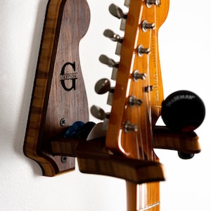 Puede incluir: Un soporte de pared de madera para guitarra con una inscripción personalizada que dice "Greyson". El soporte está hecho de madera oscura con un acento de madera clara. Una guitarra está colgada del soporte.
