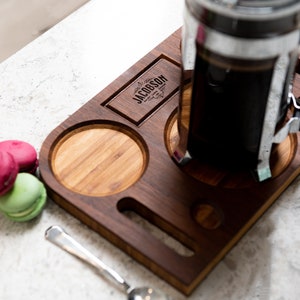May include: A wooden serving tray with a built-in holder for a French press coffee maker. The tray has a small engraved sign that reads "Jacobson" and a small spoon resting on the tray. There are also four small macarons on the surface next to the tray.