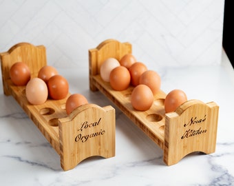 Puede incluir: Dos soportes para huevos de madera, uno etiquetado como "Local Organic" y el otro como "Nora's Kitchen". Cada soporte tiene 6 ranuras y está lleno de huevos marrones.