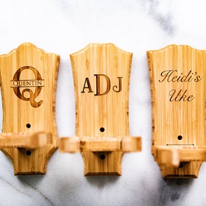 Puede incluir: Tres soportes de pared de madera para ukelele con nombres grabados. El primer soporte tiene una gran "Q" con "Quentin" escrito debajo. El segundo soporte tiene las iniciales "ADJ" grabadas. El tercer soporte tiene el texto "Heidi's Uke" grabado.