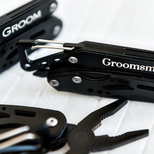Könnte beinhalten: Drei schwarze Multifunktions-Taschenmesser mit Karabinerhaken. Die Messer sind mit den Worten "Groom" und "Groomsman" graviert.