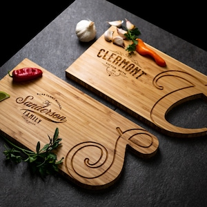 May include: Two bamboo cutting boards with engraved family names and decorative swirls. One board has "Clermont" and the other "The Sanderson Family" etched into the wood. Garnishes of garlic, peppers, and herbs are arranged on the boards.
