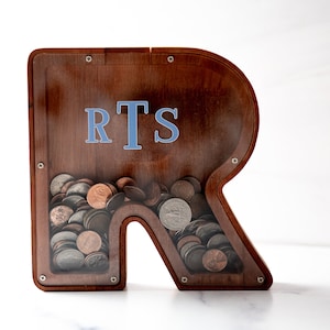 May include: A wooden piggy bank shaped like the letter R with a clear plastic front. The piggy bank has the initials "RTS" written on the front in blue letters. The piggy bank is filled with coins.