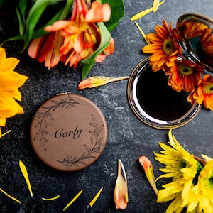 Op de afbeelding: Een ronde, bruine compactspiegel met een bloemenmotief en de naam "Carly" gegraveerd op de voorkant. De spiegel is omringd door gele en oranje bloemen.