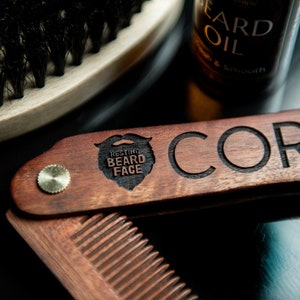 Puede incluir: Un peine de madera con un diseño plegable. El peine tiene un acento de latón y está grabado con las palabras "Resting Beard Face" y "Cory".