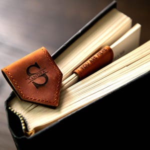 Op de afbeelding: Een bruine leren bladwijzer met de naam "Sandra" en de letter "S" erop gegraveerd. De bladwijzer ligt op een gesloten boek met de tekst "You are here" op de bladwijzer.