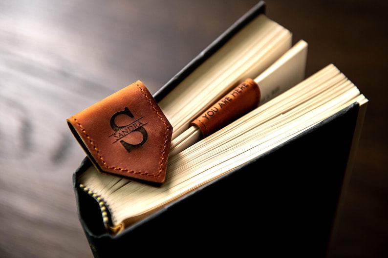 Op de afbeelding: Een bruine leren bladwijzer met de naam "Sandra" en de letter "S" erop gegraveerd. De bladwijzer ligt op een gesloten boek met de tekst "You are here" op de bladwijzer.