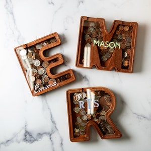 May include: Three wooden coin banks shaped like the letters E, M, and R. Each bank has a clear plastic front and is filled with coins. The letters are personalized with the names Emma, Mason, and  Rts.