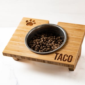 May include: A raised bamboo dog bowl stand with a stainless steel bowl filled with dog food. The stand has a paw print design and is personalised with the name "Taco".