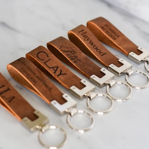 May include: Six brown leather keychains with silver key rings. The keychains are engraved with the names "JLA", "To the moon and back I love you", "CLAY", "Britt", "Haywood", and "11.28.2017 12.29.2018".