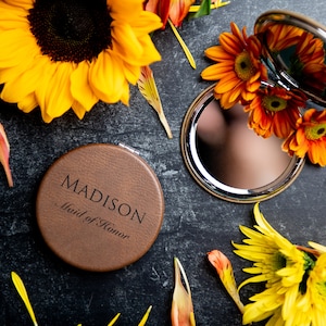 Op de afbeelding: Een compact spiegel van bruin leer met de tekst "MADISON Maid of Honor" gegraveerd op de voorkant. De spiegel is omringd door gele en oranje bloemblaadjes.