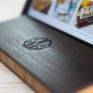 May include: A dark wooden box with a light wood trim and a hexagonal logo engraved on the top. The logo is a stylized design of the letters "B" and "K".